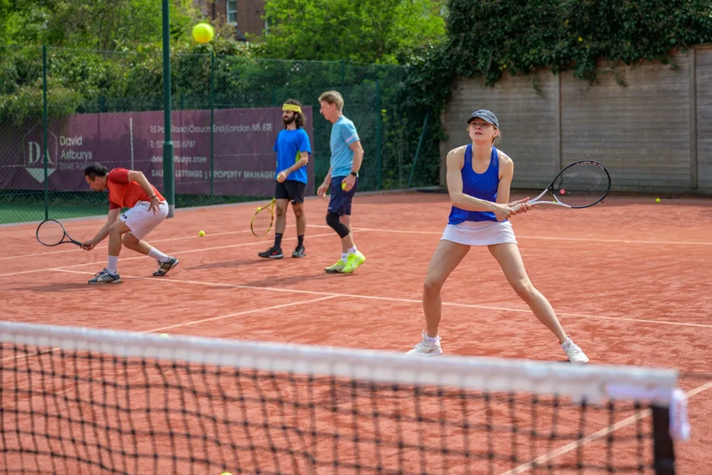 Group of tennis players on a red clay court at Coolhurst Tennis Club. One woman in blue and white is ready to return a shot at the front, while others prepare in the background near the fence and net.