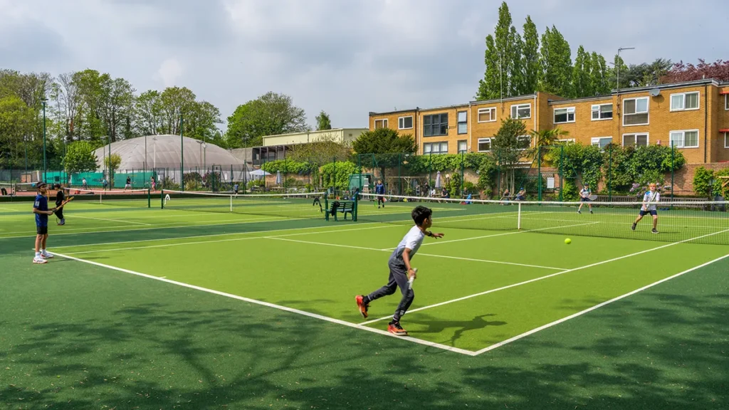 Children play tennis on a green hard court at Coolhurst Tennis Club, with residential buildings and trees visible in the background on a partly cloudy day.