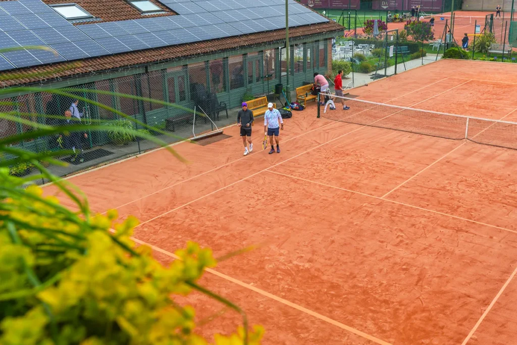 Two men walk along a red clay tennis court beside a clubhouse with solar panels, while other players are visible on adjacent courts surrounded by greenery.