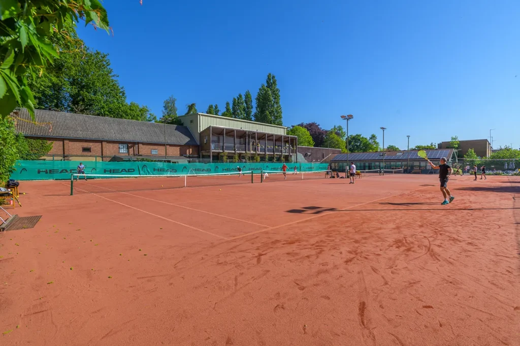 Outdoor clay tennis courts at Coolhurst Tennis Club on a bright sunny day with players practicing and a clubhouse visible in the background.